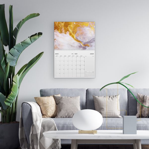 Sieninis kalendorius, sofa ir trys pagalvės ant jos, šalia sofos tovi gėlė dideliais lapais ir priešais staliukas su lempa.
